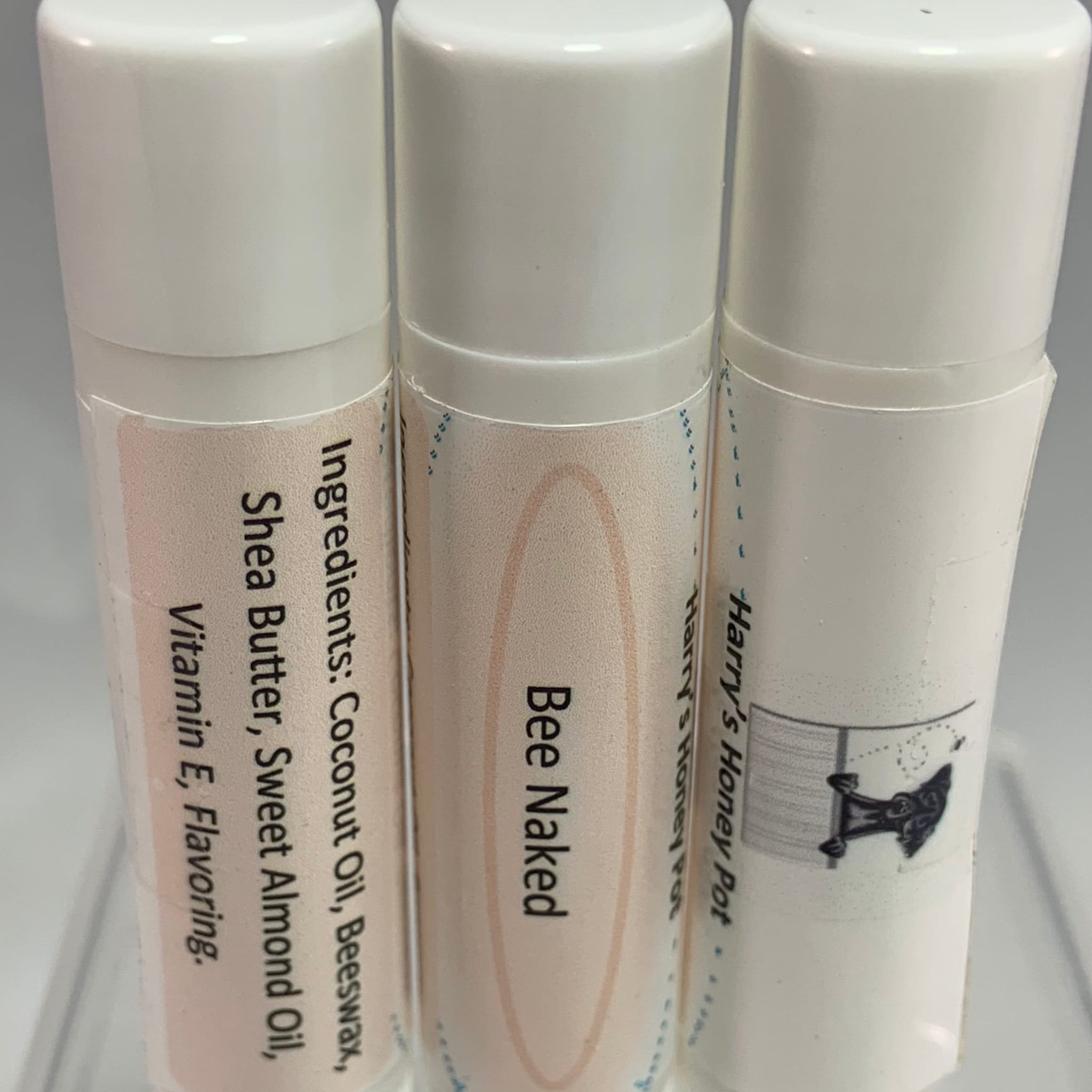 Lip balm tubes with white caps, light pink labels, and text including "Bee Naked" and "Harry's Honey Pot