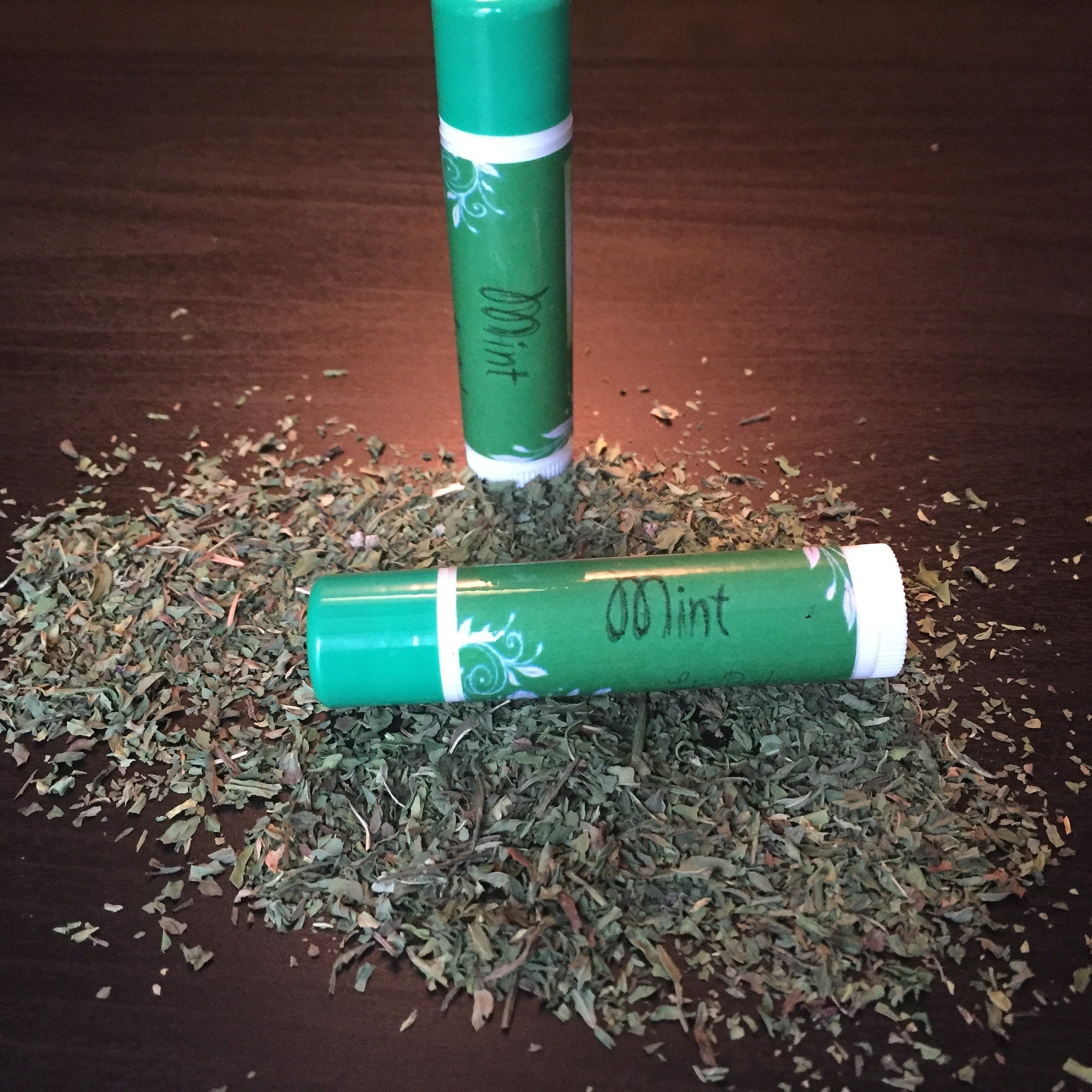 Lip balm tubes, green with white accents and "Mint" text, on dried herbs
