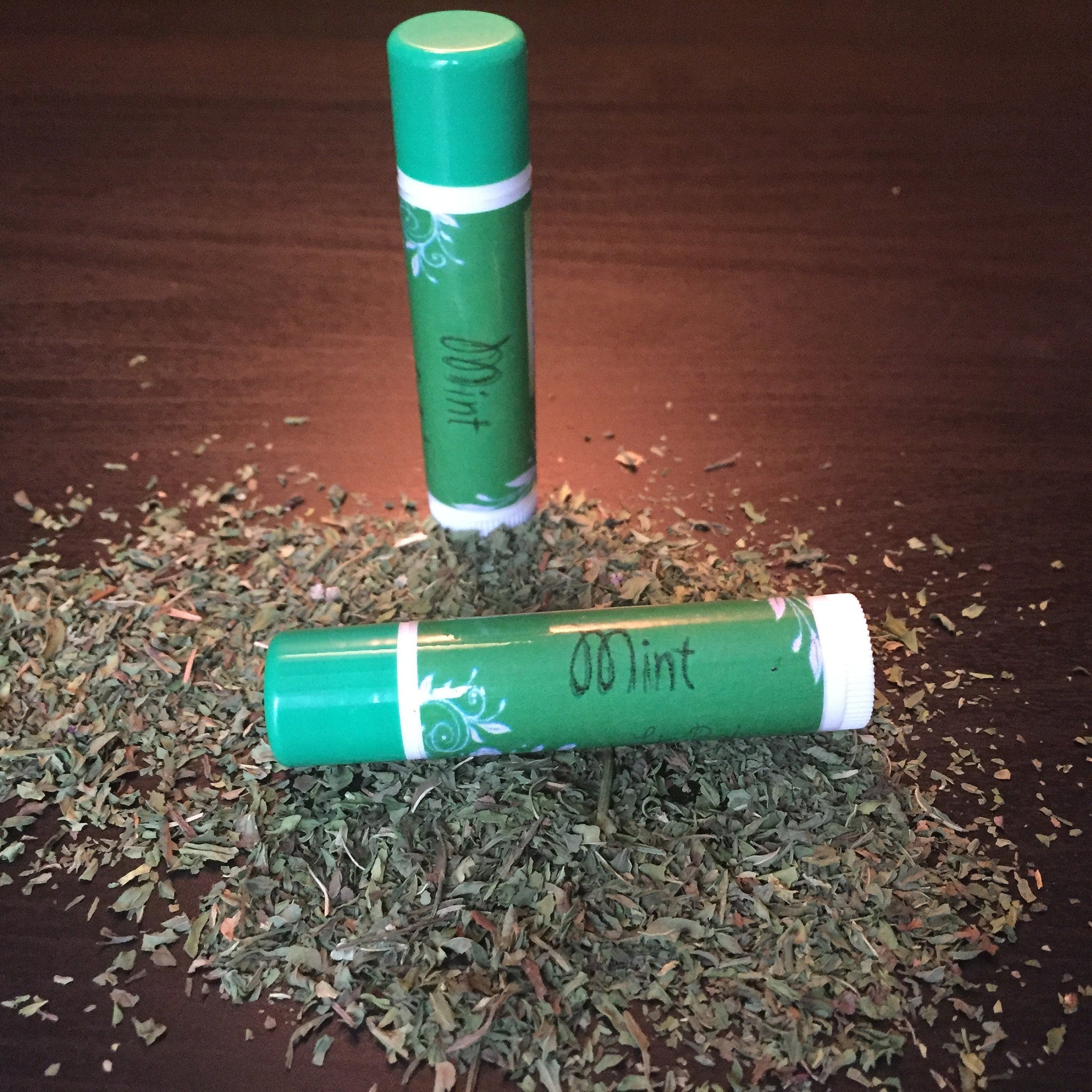 Lip balm tubes, green and white, with "Mint" text, on a bed of herbs