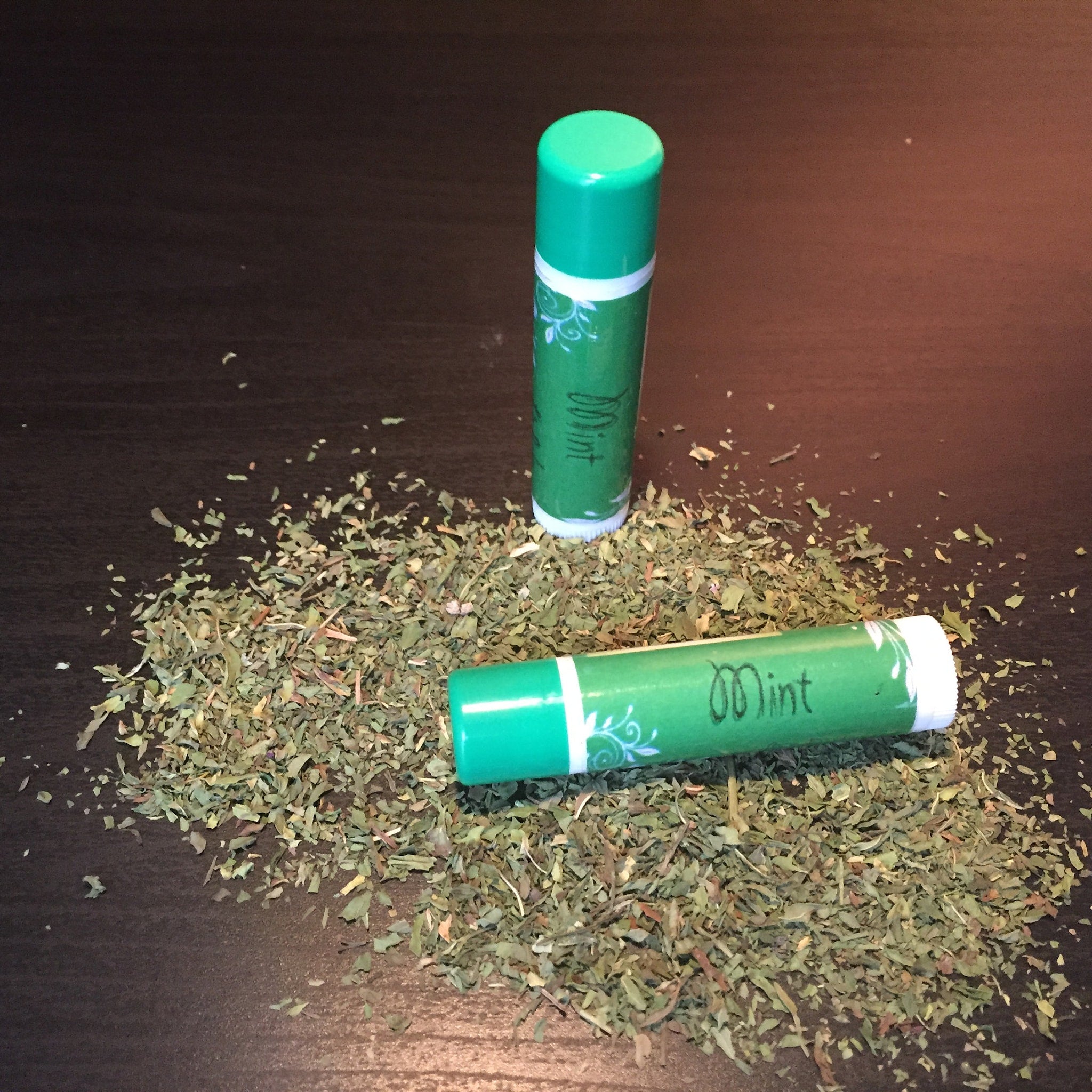 Lip balm tubes with green and white labels, "Mint" text, and dried mint leaves on surface