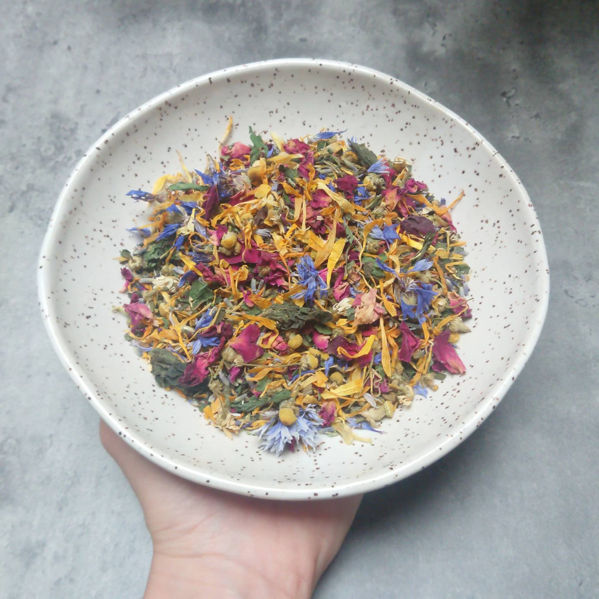 Herbal tea blend with colorful dried flowers in a speckled ceramic bowl held by a hand