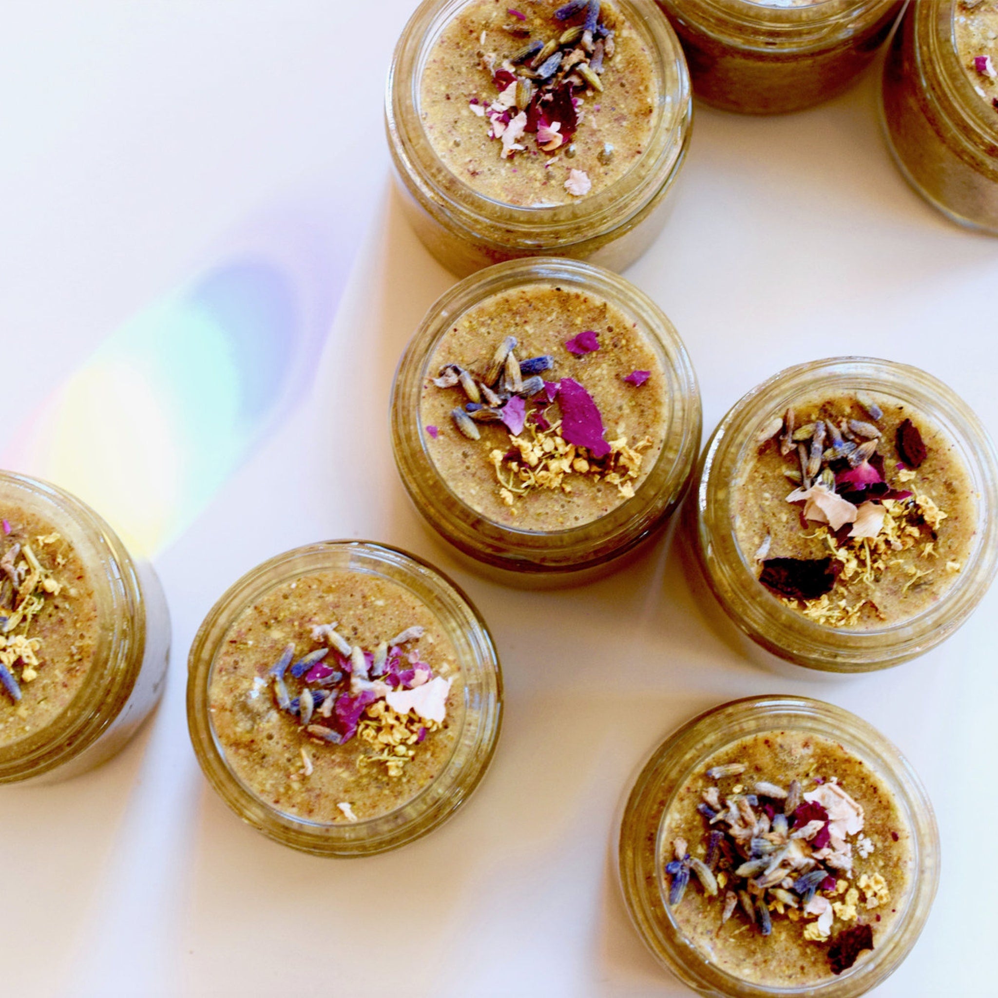 Body scrub jars with brown scrub, dried flowers, and glass jars on white surface