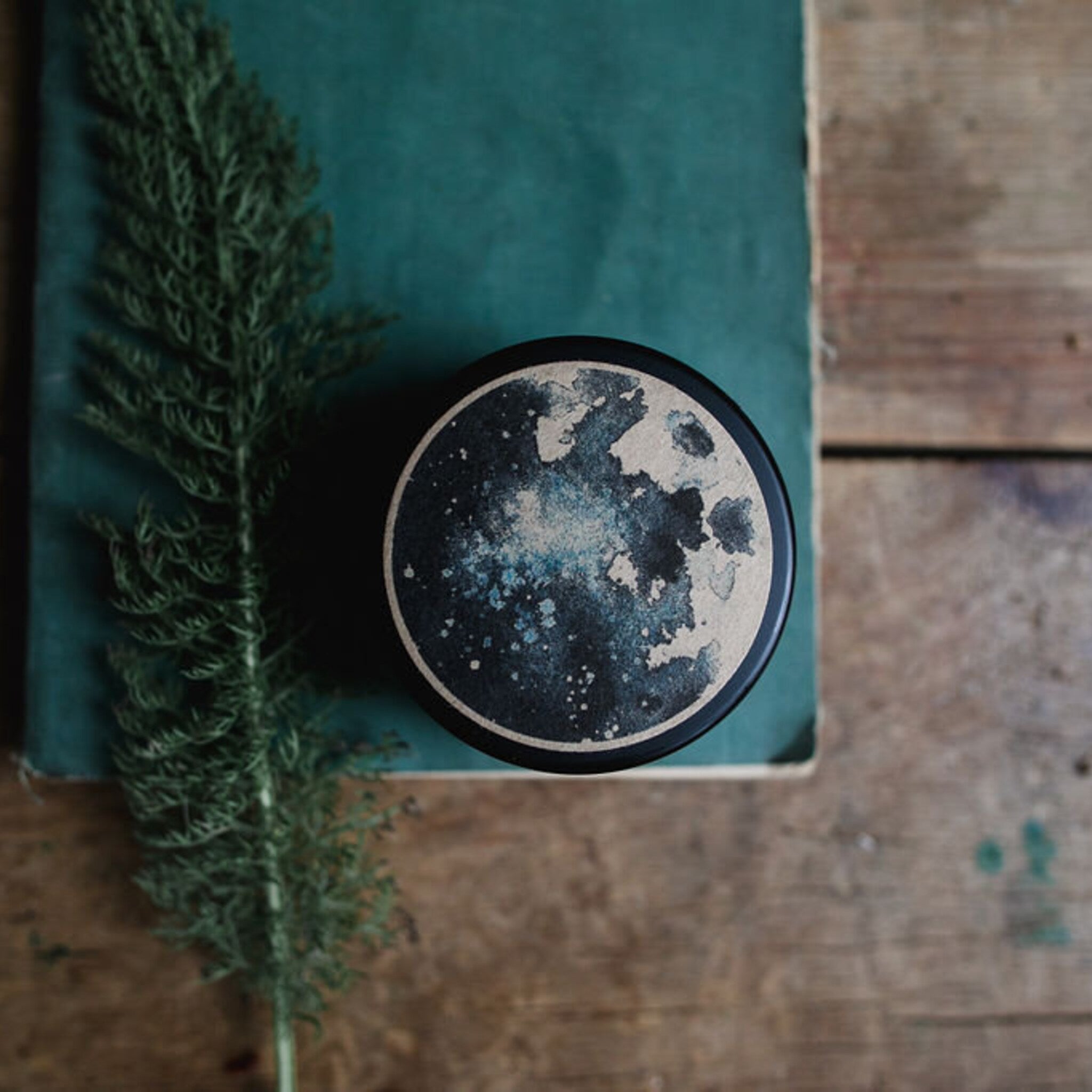 Round black container with moon design, on green paper and wooden surface, with green plant