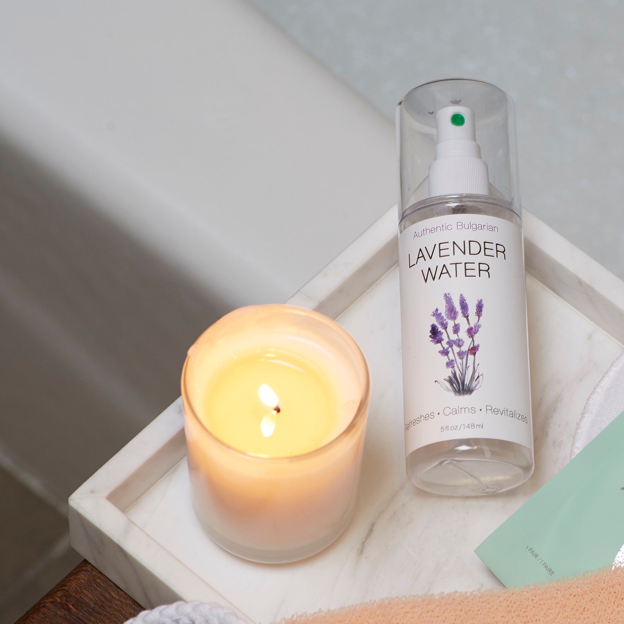 Lavender water spray bottle with white label, purple floral illustration, and clear plastic top, next to lit candle on marble tray