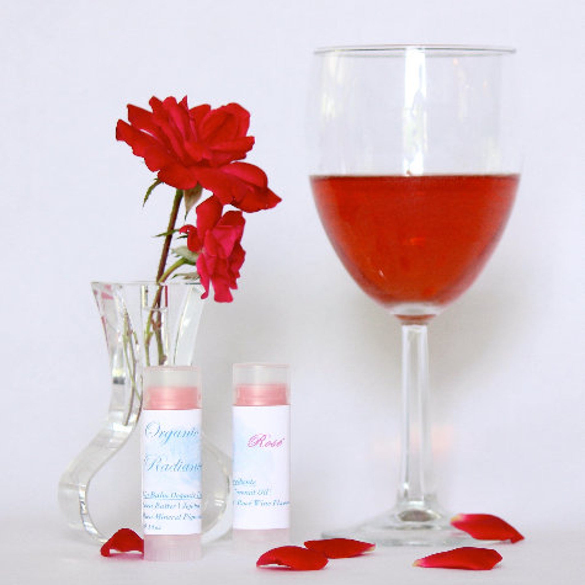 Lip balm tubes with white labels, red rose petals, and a glass of red liquid on white background