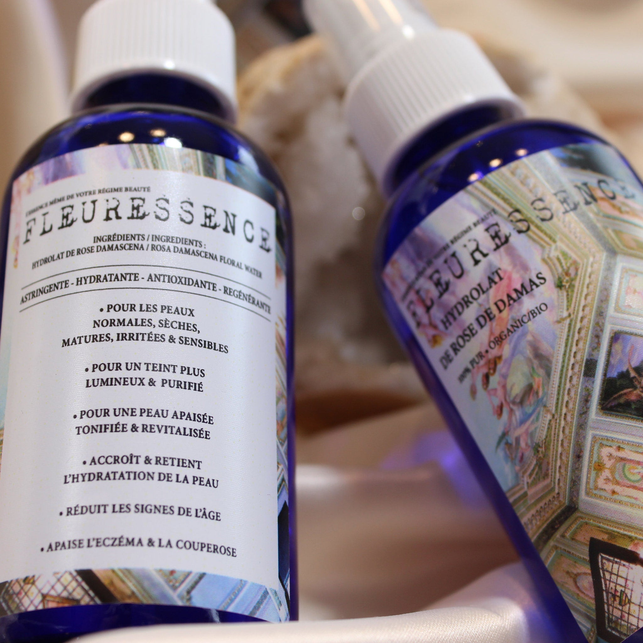 Floral water bottles, blue glass with white sprayers, FLEURESSENCE label visible, on white fabric surface