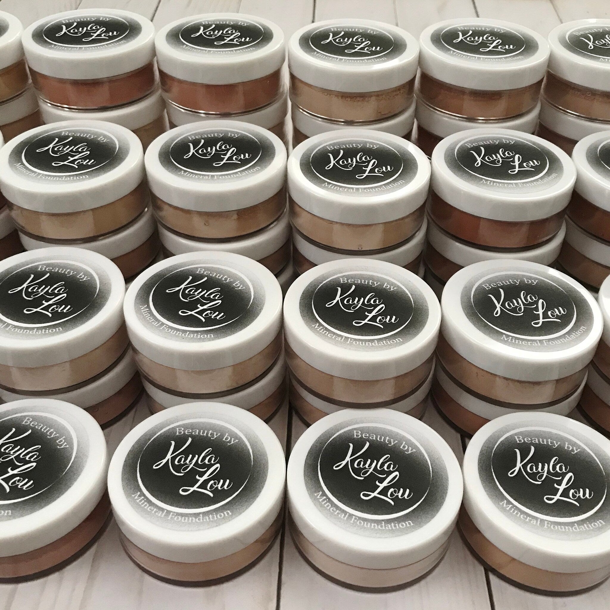 Mineral foundation jars with white lids, various shades of powder, and "Beauty by Kayla Lou" label