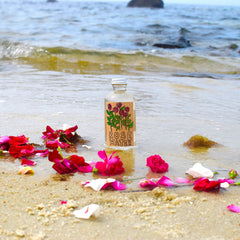 Rose water bottle with clear glass, silver cap, and "Rose Water" label on beach with petals