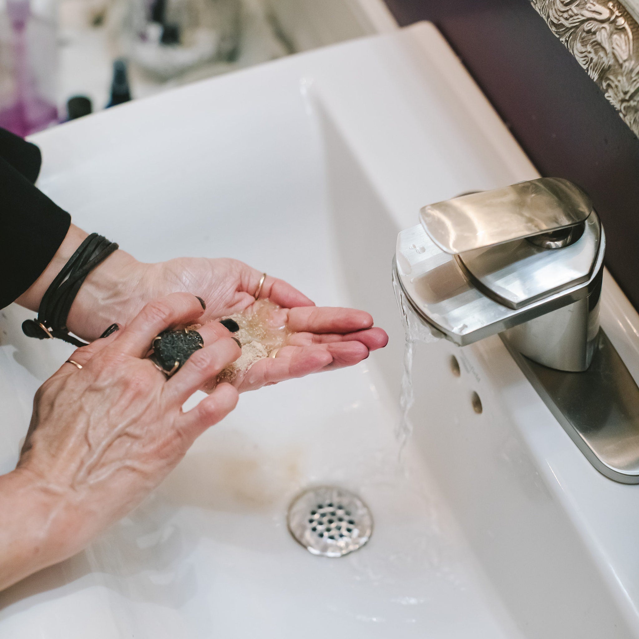Hands washing with clear gel soap, black nail polish, gold rings, and black leather bracelet in white sink