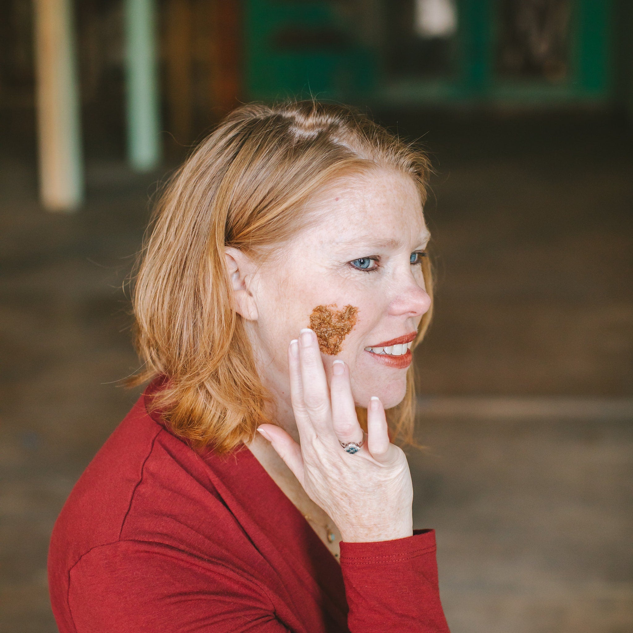 Woman with red shirt and light brown hair, applying brown mask to face