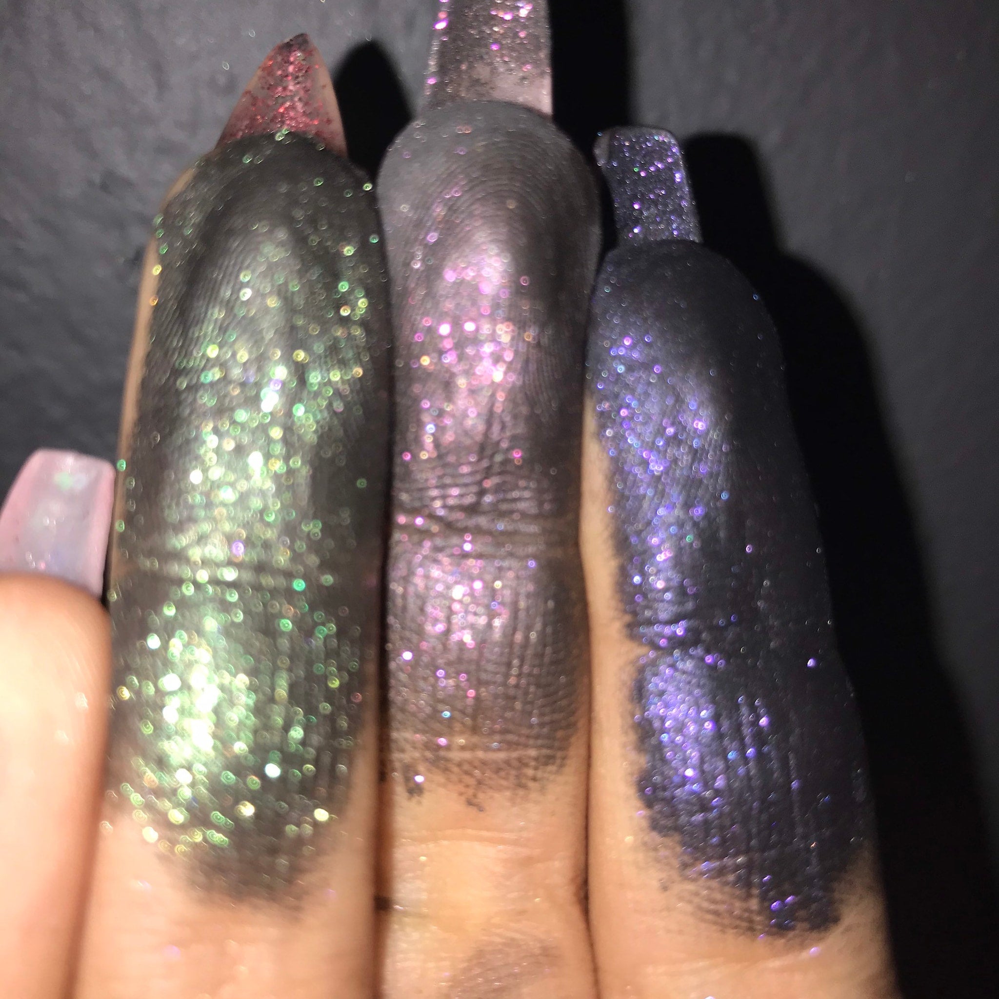 Glitter eyeshadow swatches in green, pink, and purple on fingers, close-up view against a dark background