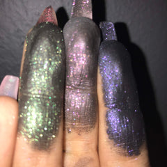 Glitter eyeshadow swatches in green, pink, and purple on fingers, close-up view against a dark background