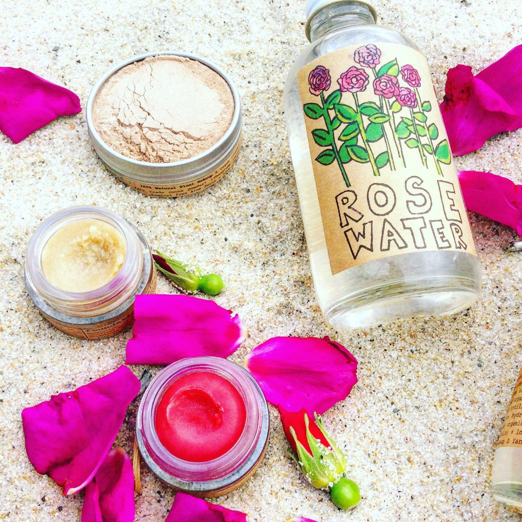 Rose water bottle with label, pink rose petals, and lip balm on sand