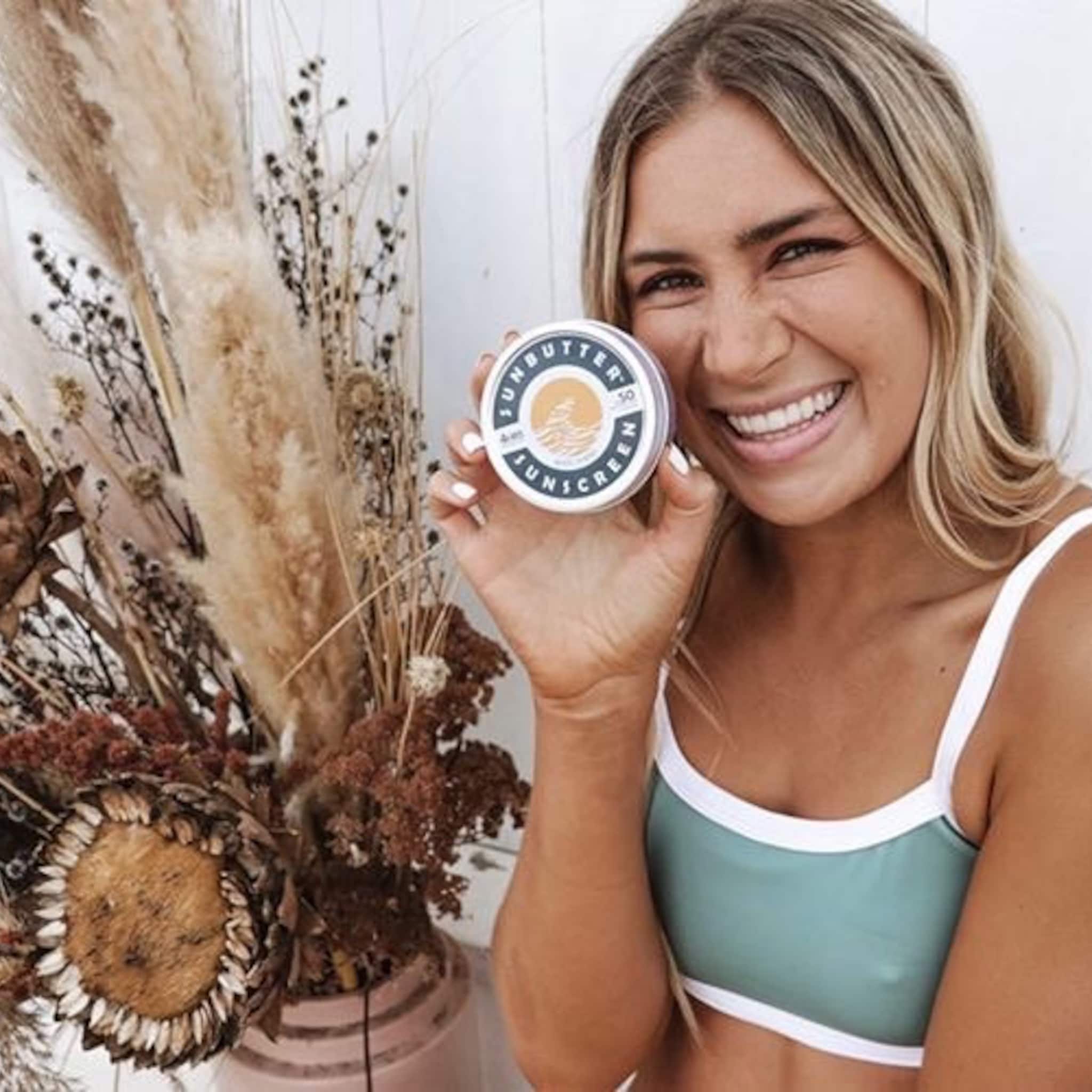 Sunscreen tin with white and blue text held by smiling woman in green top
