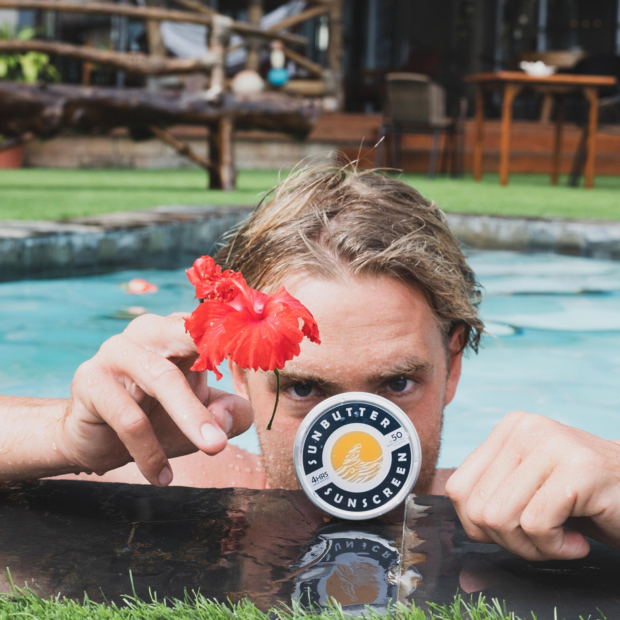 Sunscreen tin with white and blue label, held by man near pool with red flower