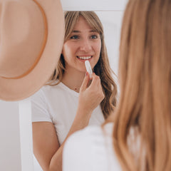Lip balm white tube with text, being applied by woman in white shirt, reflected in mirror
