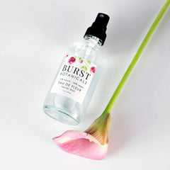Facial mist bottle with black sprayer, clear glass, and "BURST BOTANICALS" label with flower pattern, next to pink calla lily