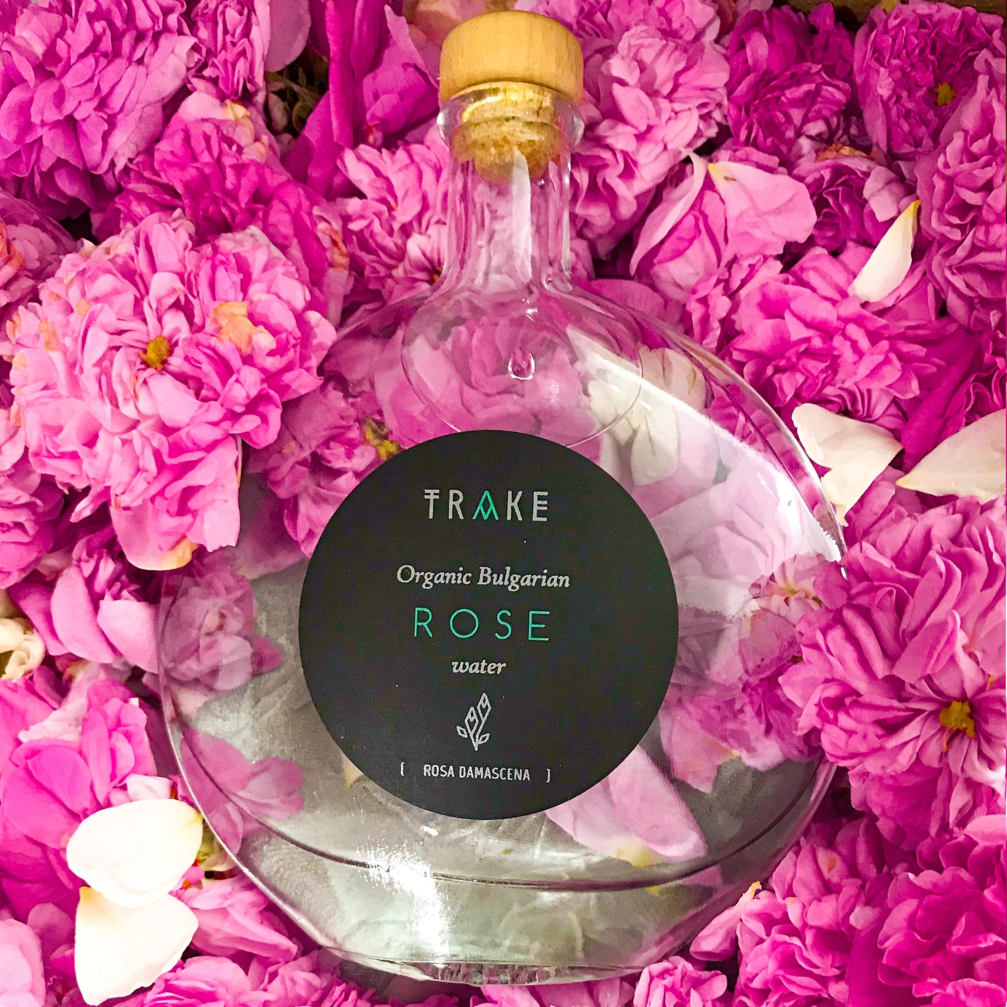 Rose water bottle with glass, wood, and black label, surrounded by pink flowers, TRAKE brand visible