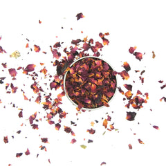 Dried rose petals in a metal bowl, with red, orange, and brown hues scattered on white surface