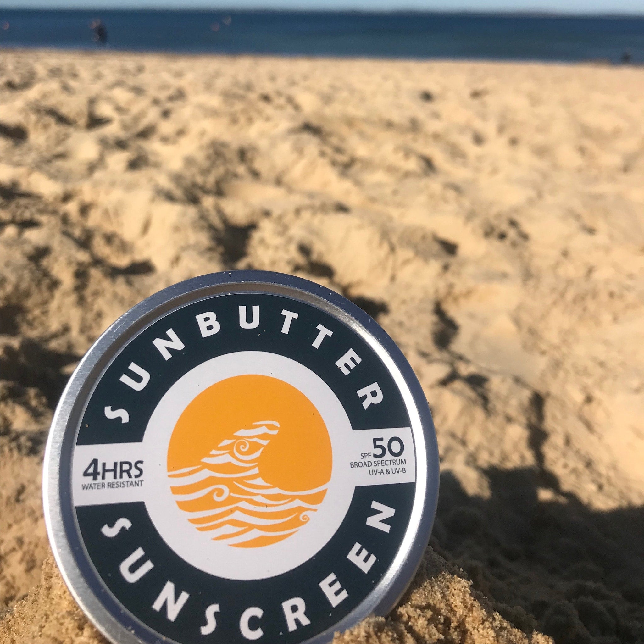Sunscreen tin with black, white, and orange design on sand near ocean