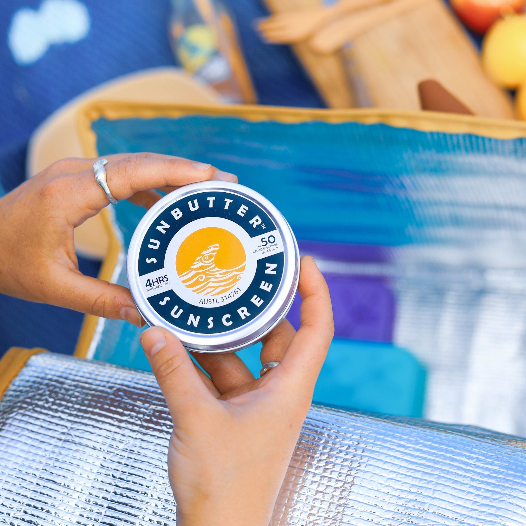 Sunscreen tin with blue and white label, held by hands, on a silver insulated surface, SUN BUTTER text visible