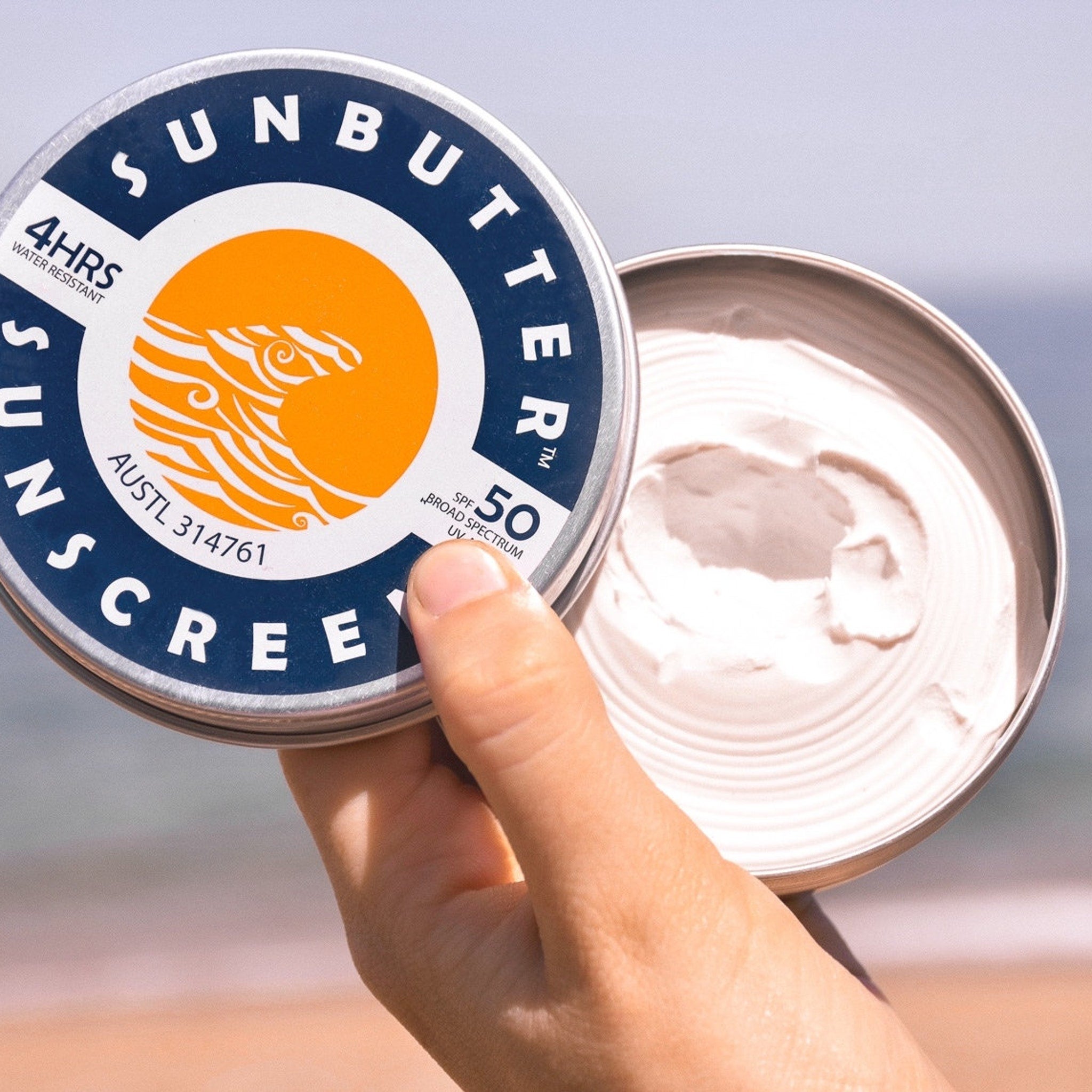 Sunscreen tin with blue and white text, orange logo, and cream-colored cream inside, held by hand