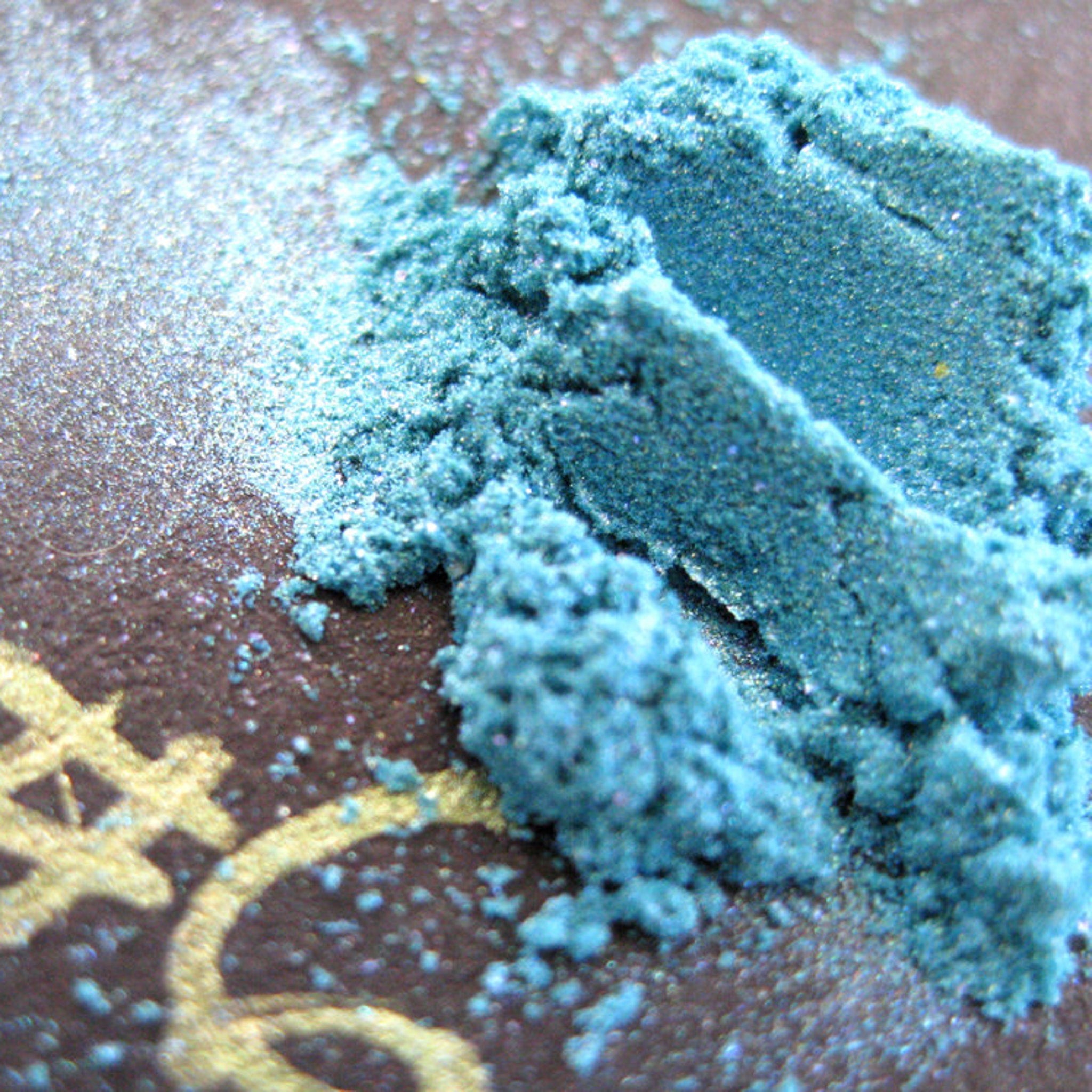 Turquoise blue loose eye shadow in a clear jar with iridescent aqua and purple shimmer particles visible.