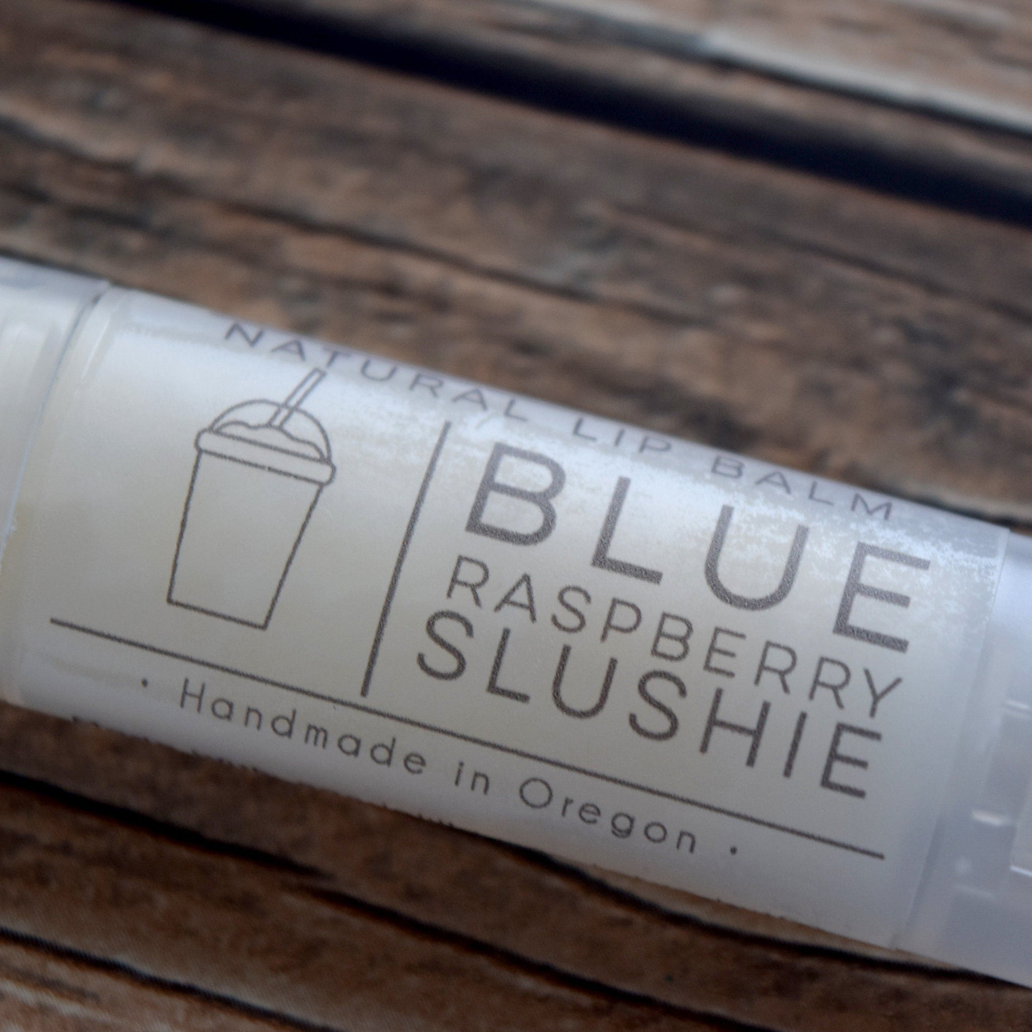 Lip balm tube with white label, gray text "Blue Raspberry Slushie", and a cup graphic on a wooden surface