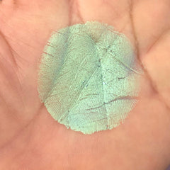 Green pigment swatch on skin, round shape, textured appearance, hand in background