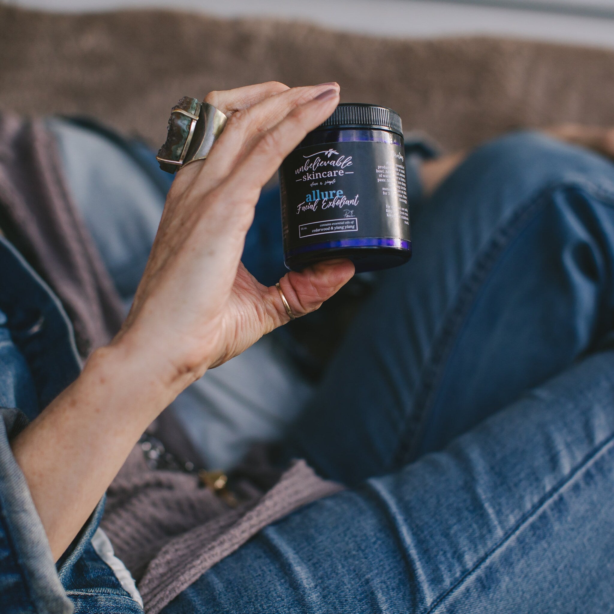 Facial Exfoliant jar with black lid, blue label, and text held in hand with jewelry.
