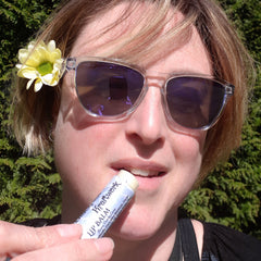 Lip balm tube with white and blue label, being applied to lips by person wearing sunglasses