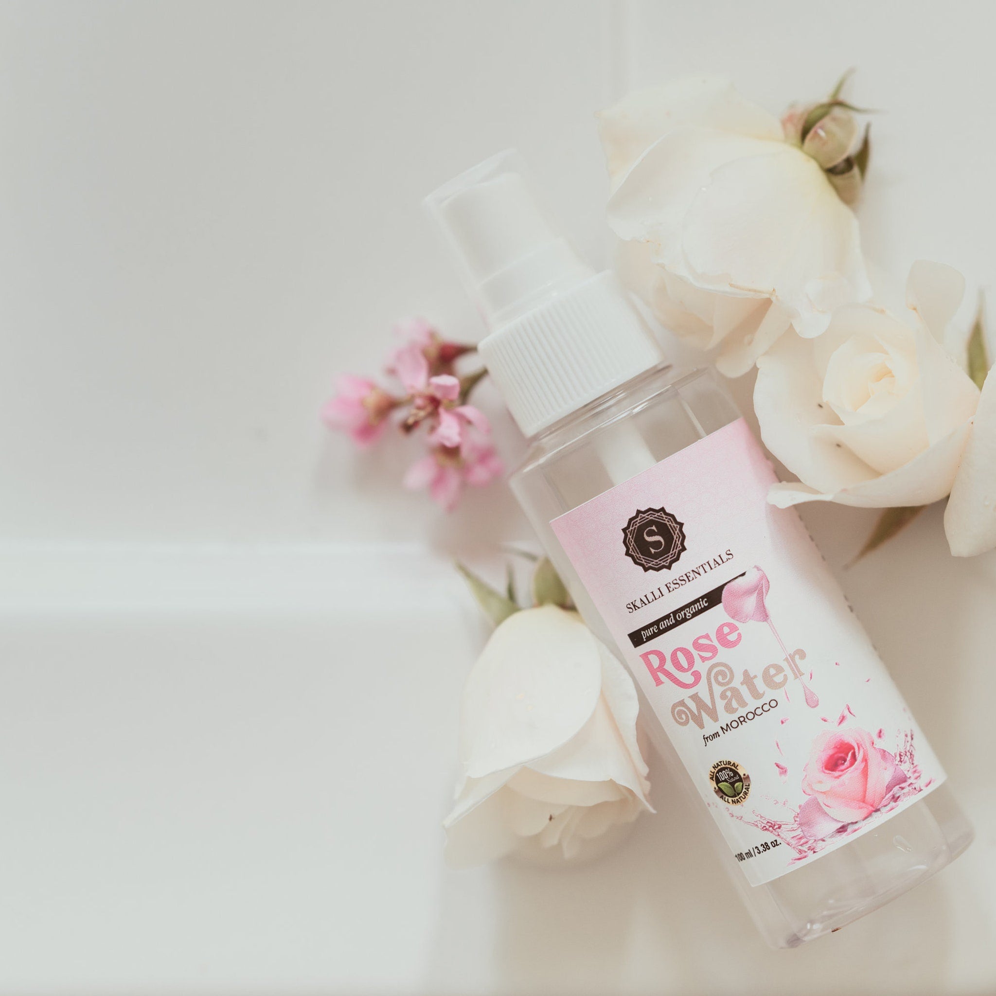 Rose water spray bottle with white and pink label, surrounded by white roses and pink flowers