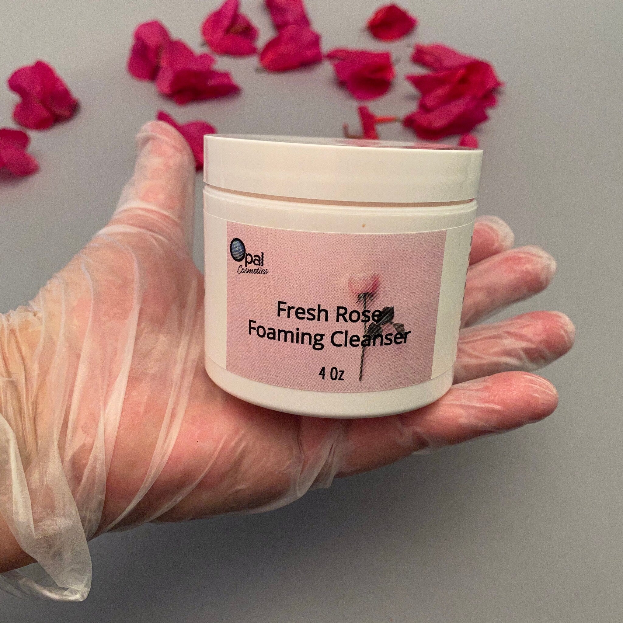 Foaming cleanser jar, white container with pink label, held in gloved hand with pink flowers in background