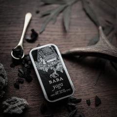 Lip balm tin in black with Baba Yaga text, silver spoon, and wooden background