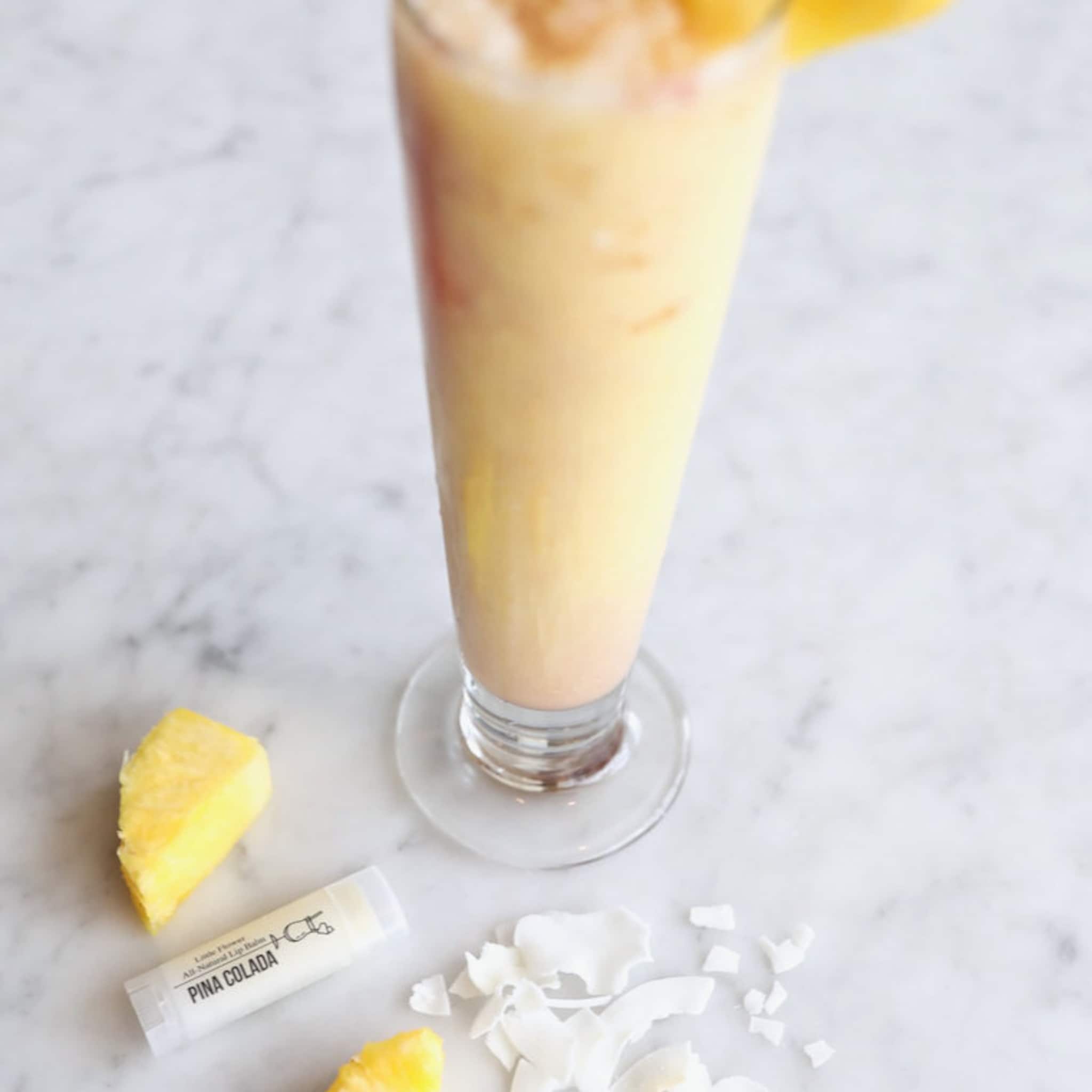 Pina Colada lip balm tube, white and clear, with yellow pineapple and coconut flakes on marble surface