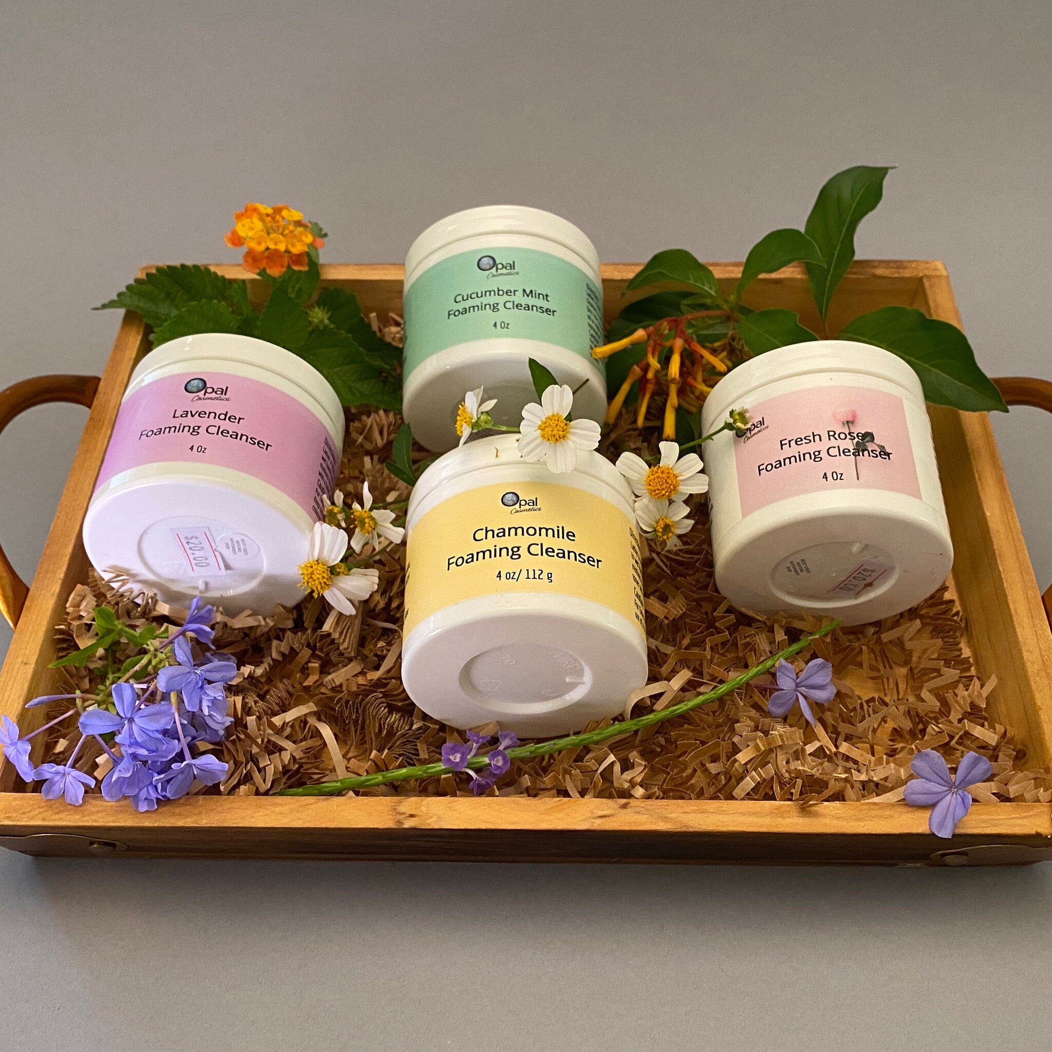 Foaming cleanser jars in white, with colorful labels, displayed in a wooden tray with flowers and shredded paper