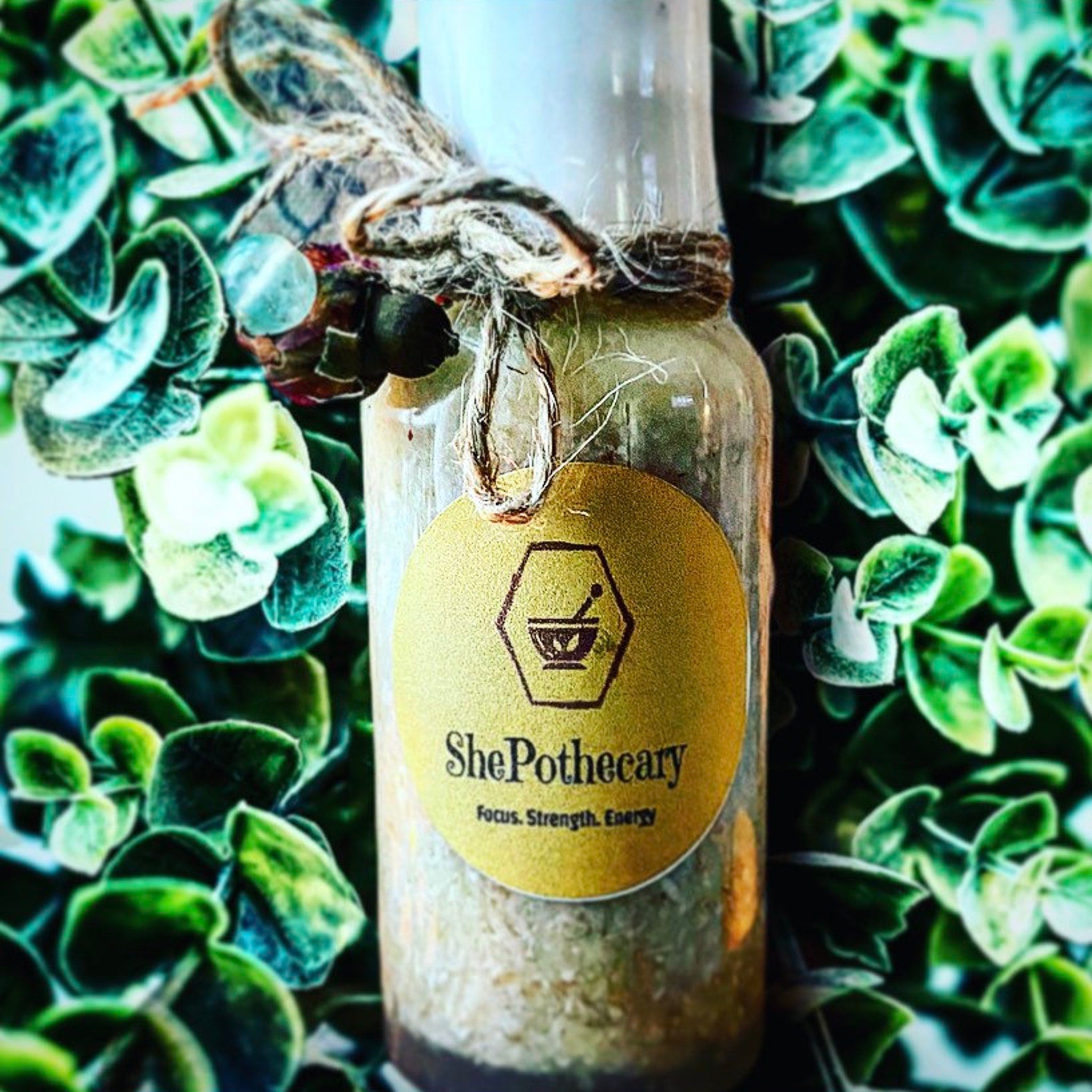 ShePothecary bottle with gold label, twine tie, and green foliage background
