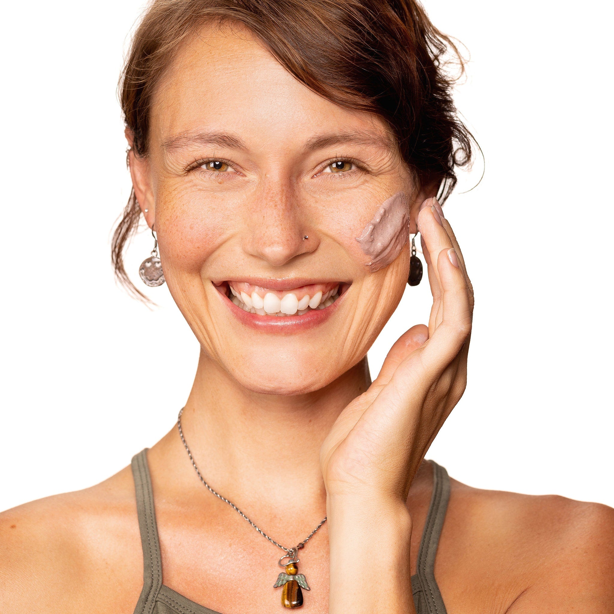 Woman applying face mask, smiling with brown hair and green tank top, with jewelry on