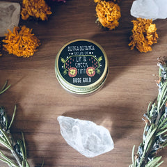 Lip and cheek tint Rose Gold in a black and gold tin, surrounded by flowers and crystals on wood