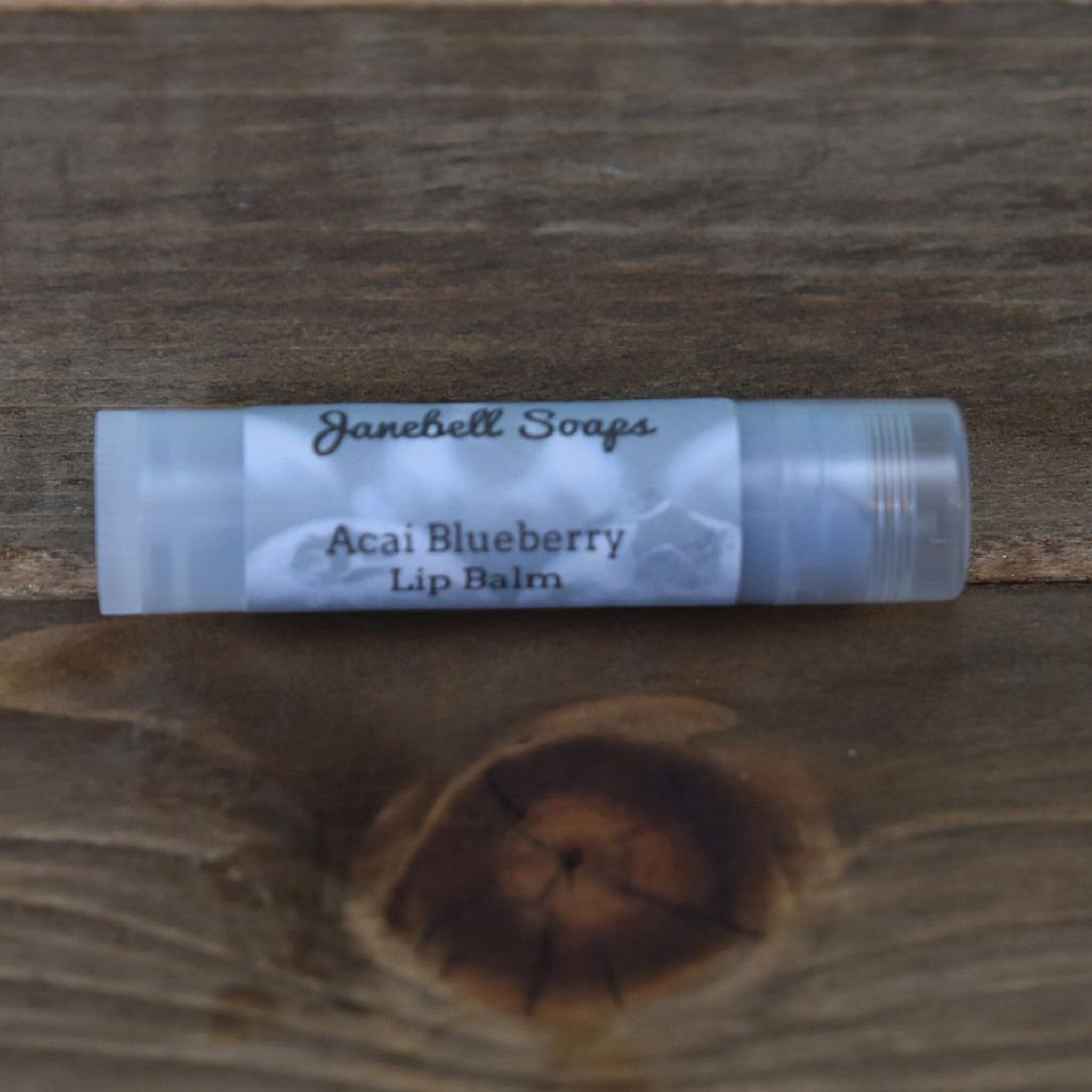 Lip balm tube with white label, clear plastic, and Janebell Soaps text on wooden surface