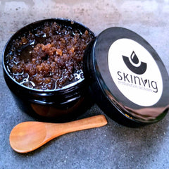 Brown scrub in black jar with Skinvig label and wooden spoon on gray surface