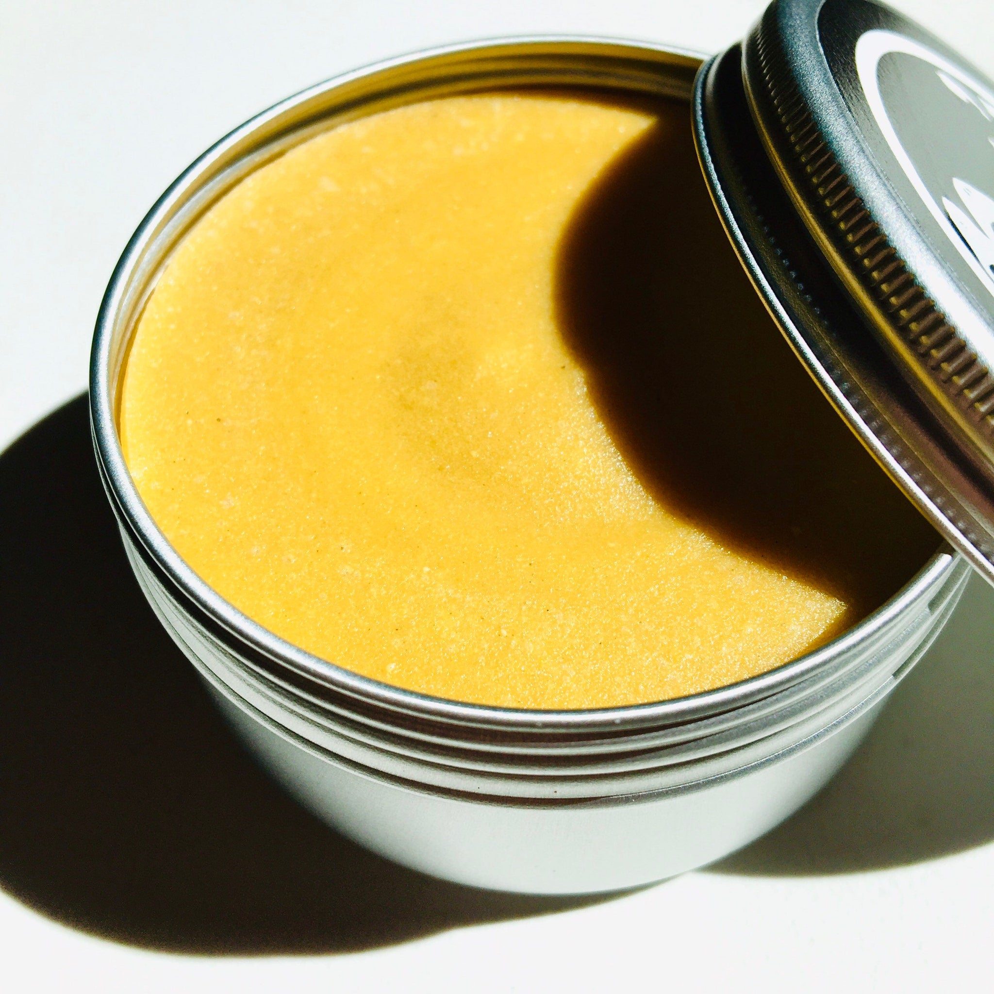 Yellow balm in a silver metal tin with a lid partially open, close up.