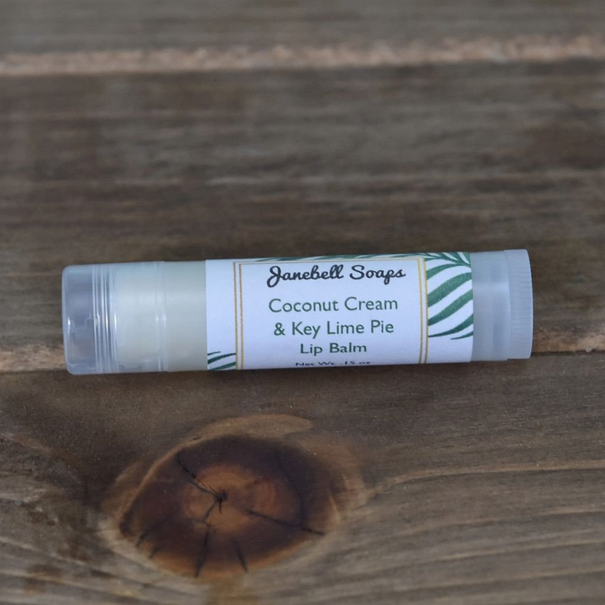 Lip balm tube with white label, clear plastic, and Janebell Soaps branding on wood background