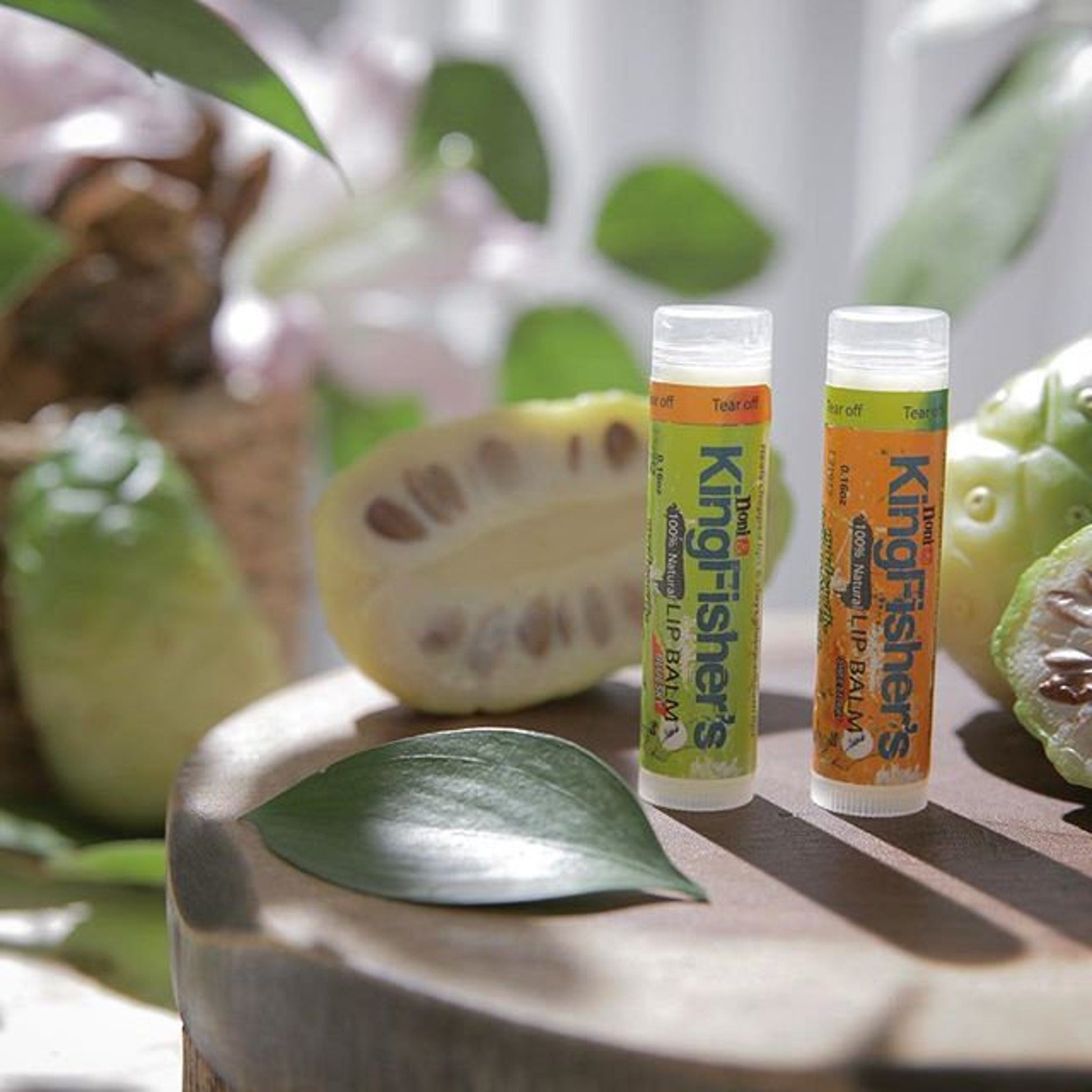 Lip balm tubes with green and orange labels, on a wooden surface with fruit and leaves, KingFisher's brand visible