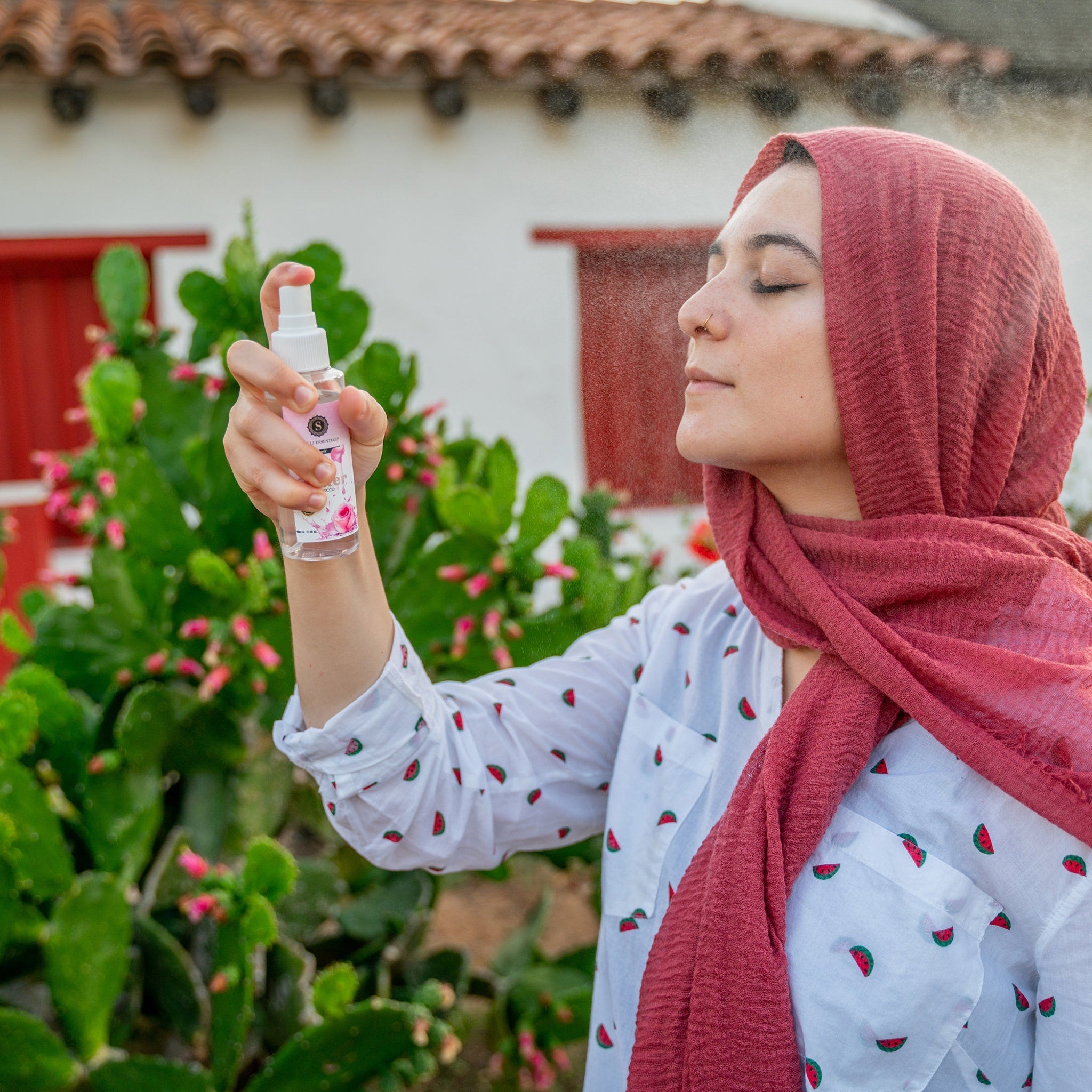 Facial mist bottle held by person, clear plastic, white spray nozzle, pink label, and red scarf