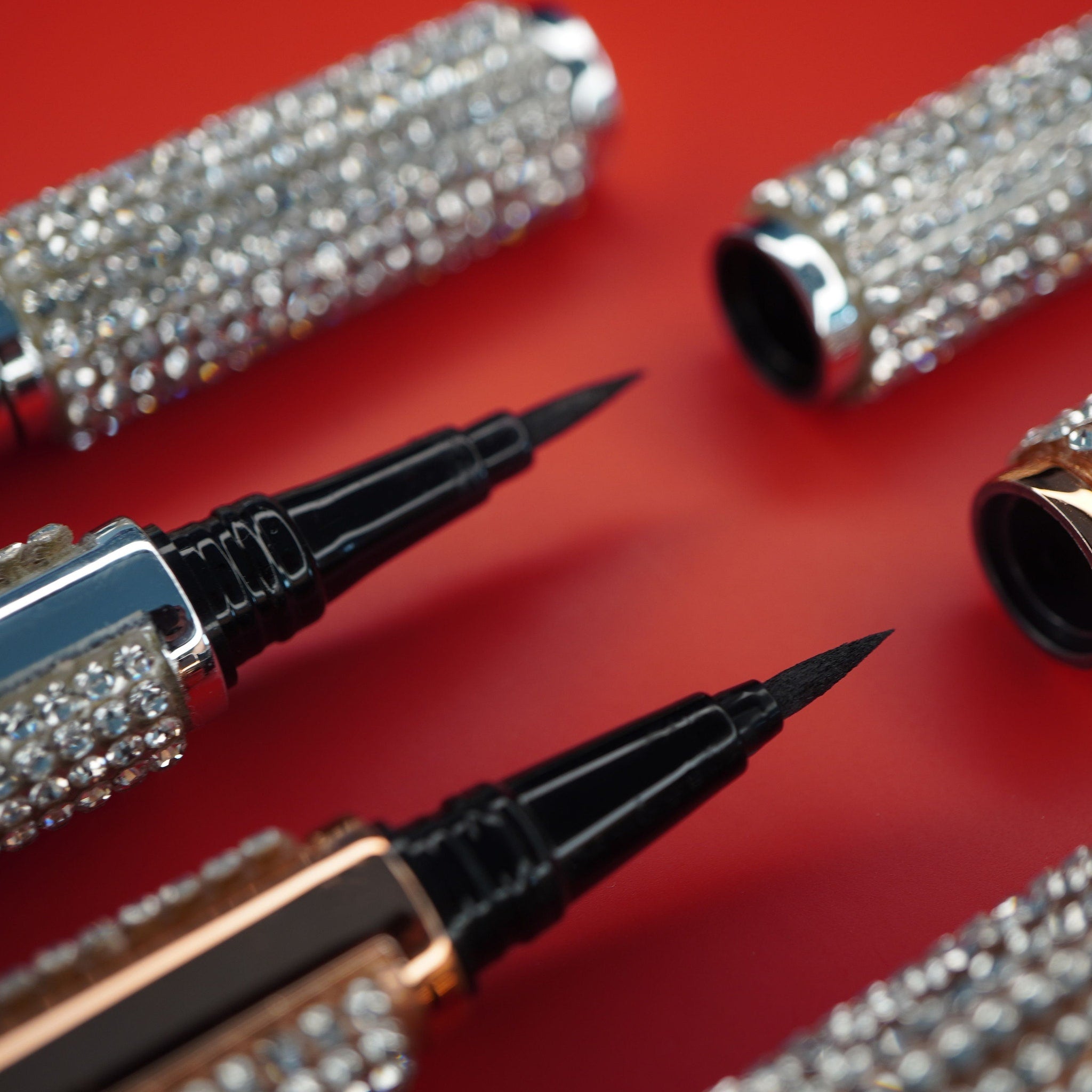 Eyeliner pens with black tips and rhinestone embellished silver and gold barrels on red surface