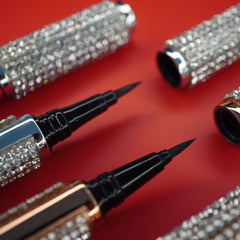 Eyeliner pens with black tips and rhinestone embellished silver and gold barrels on red surface