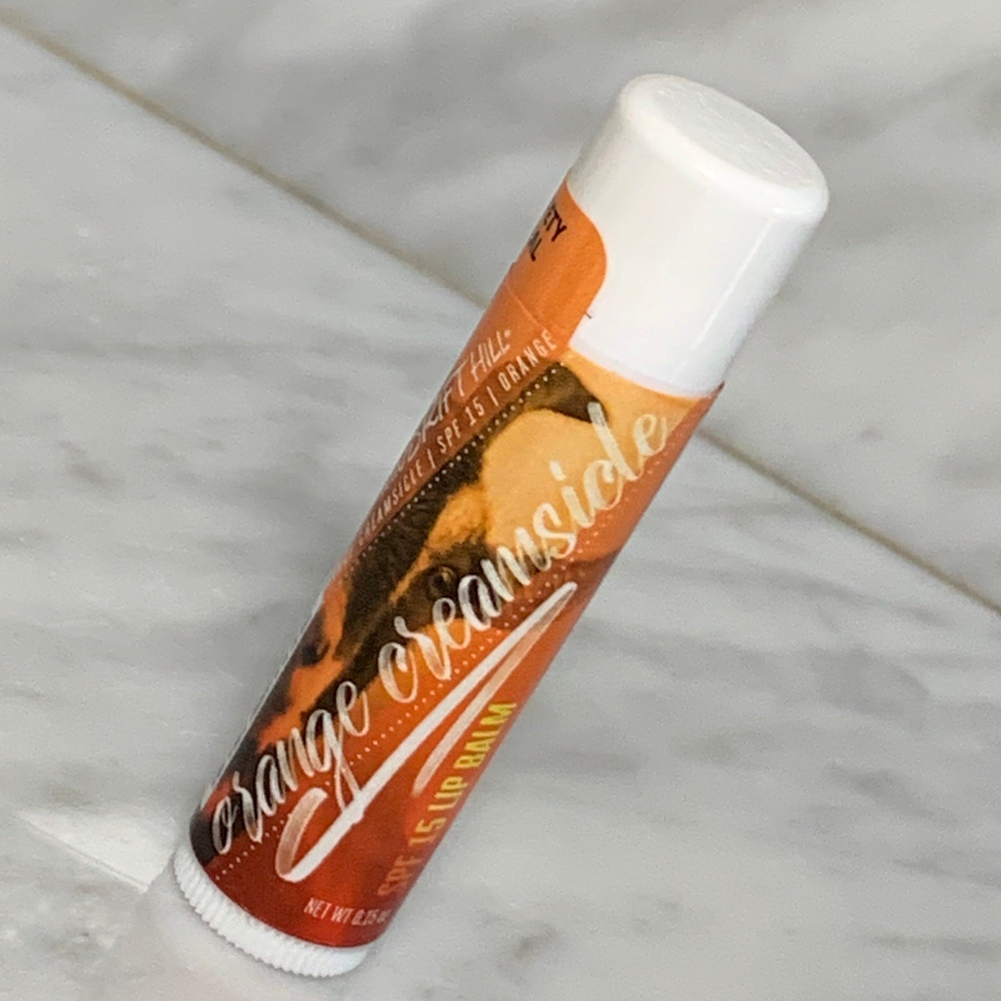 Lip balm tube with orange, white, and brown label, "Orange Creamsicle" text visible.