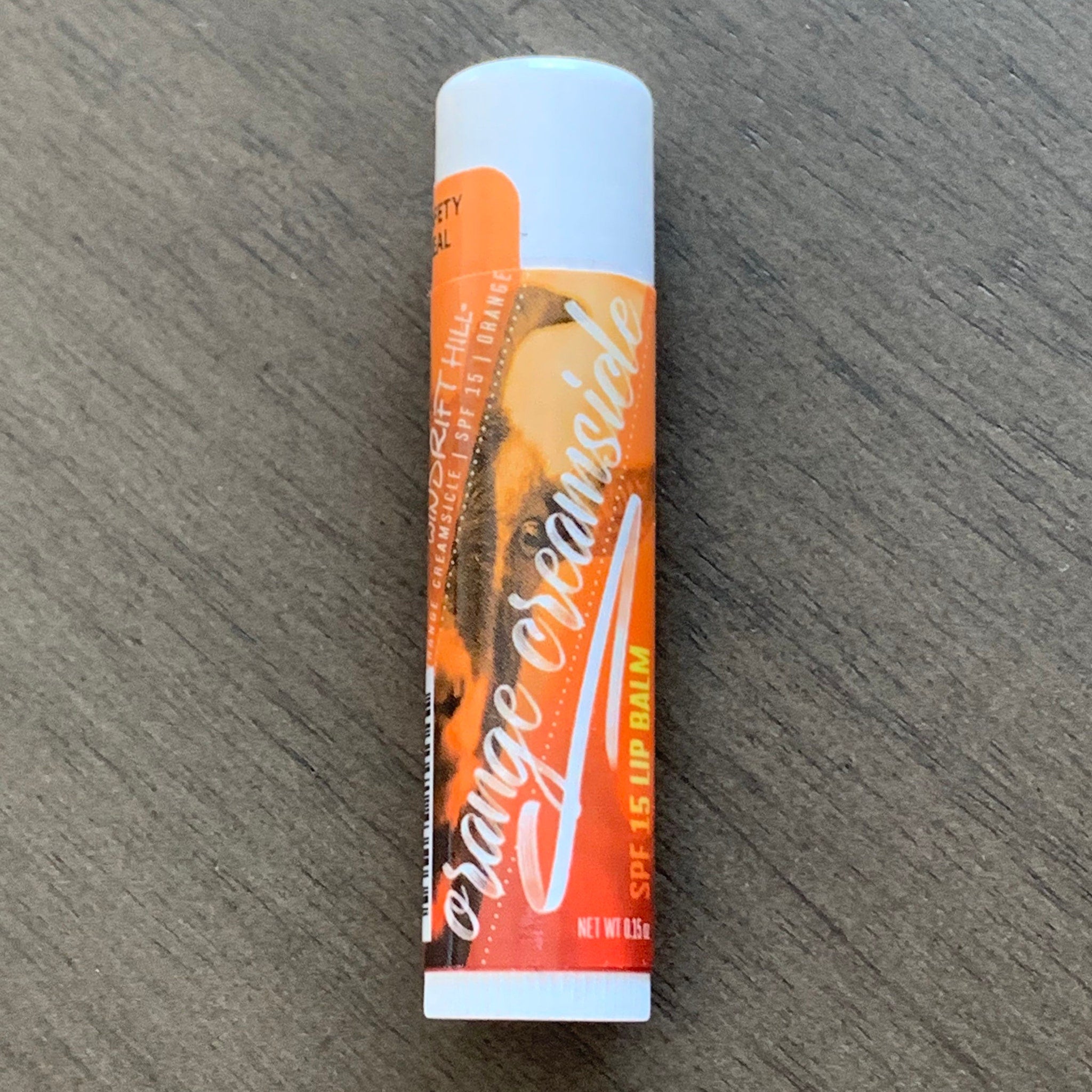 Lip balm tube with orange and white label, "Orange Creamsicle" text, on wood grain surface
