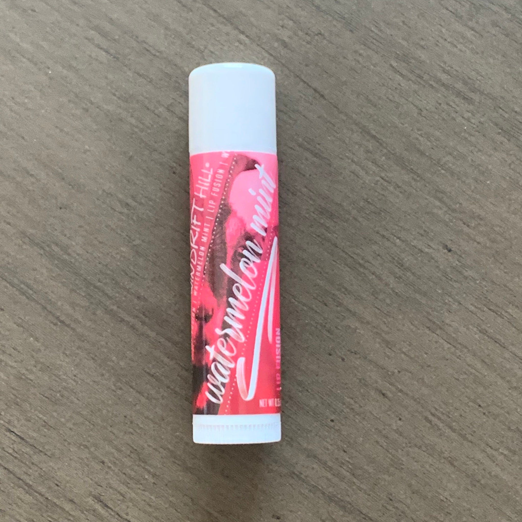 Lip balm tube with white cap, pink and red label with "Watermelon Mint" text on wooden surface